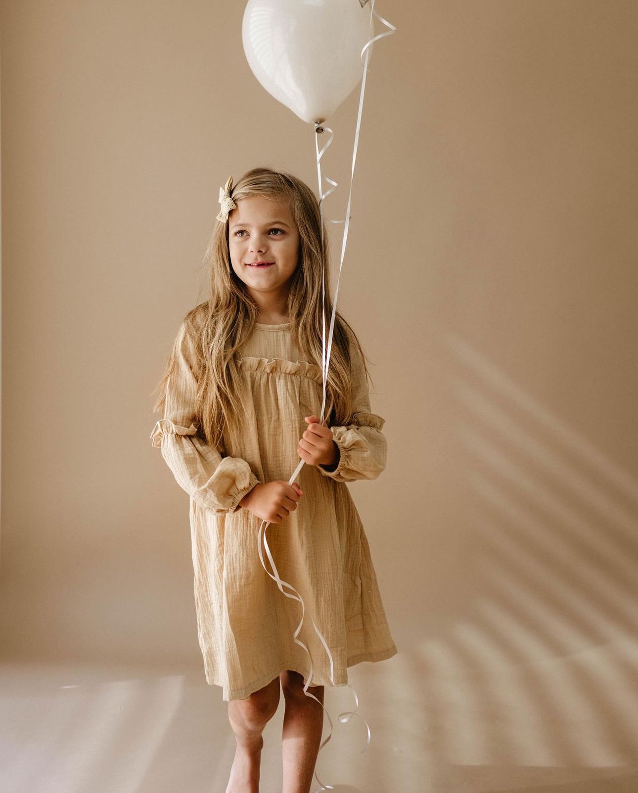 A young girl with long hair and a bow stands barefoot against a neutral background, wearing the "Golden Hour" dress by forever french baby. She holds a white balloon, and the soft light casts gentle shadows on the wall.