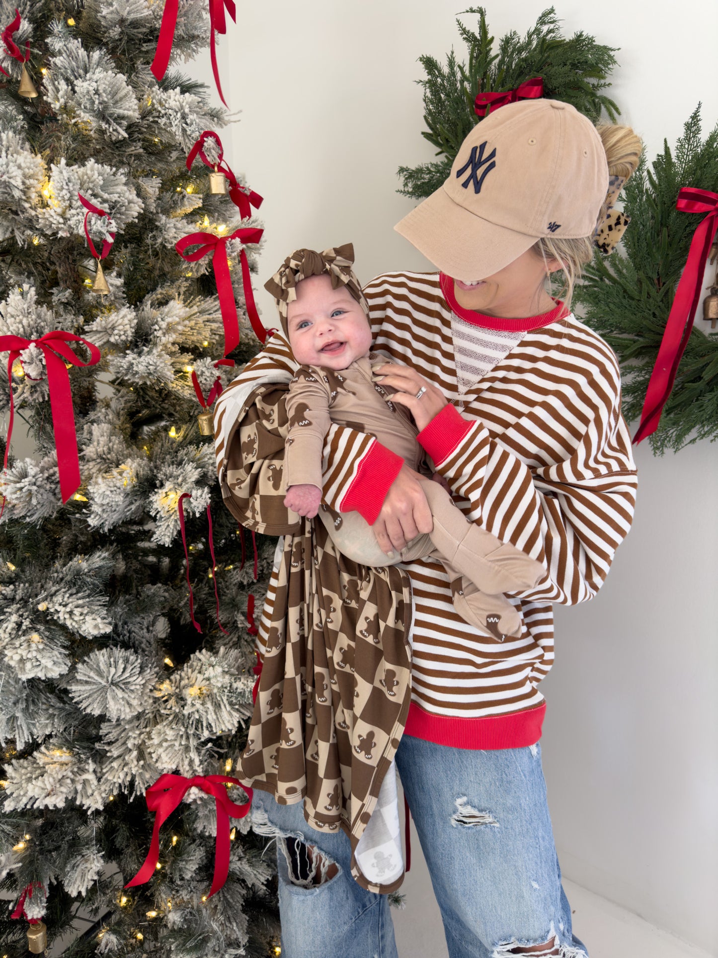 A person in a striped sweater and baseball cap holds a smiling baby dressed in a brown outfit with an adorable adjustable Bamboo Head Wrap in Gingerbread Checkerboard by forever french baby. They stand beside a Christmas tree adorned with red ribbons, positioned in front of a wreath on a white wall.