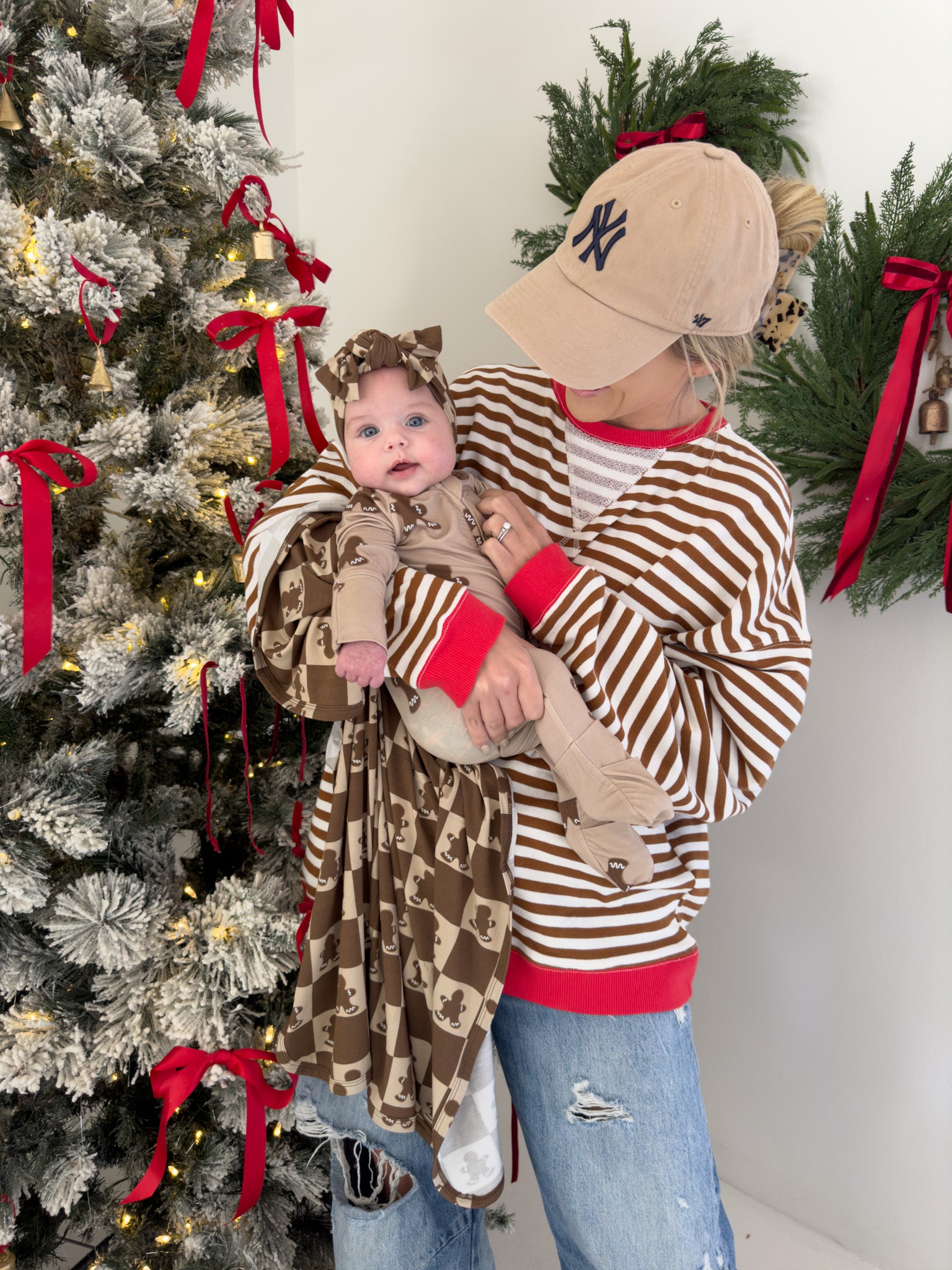 A person in a striped sweater and baseball cap holds a baby dressed in the enchanting Gingerbread Checkerboard outfit from forever french baby, crafted entirely from soft bamboo material. They stand by a beautifully decorated Christmas tree adorned with red bows and a wreath in the background, creating an ideal scene for babies and toddlers.