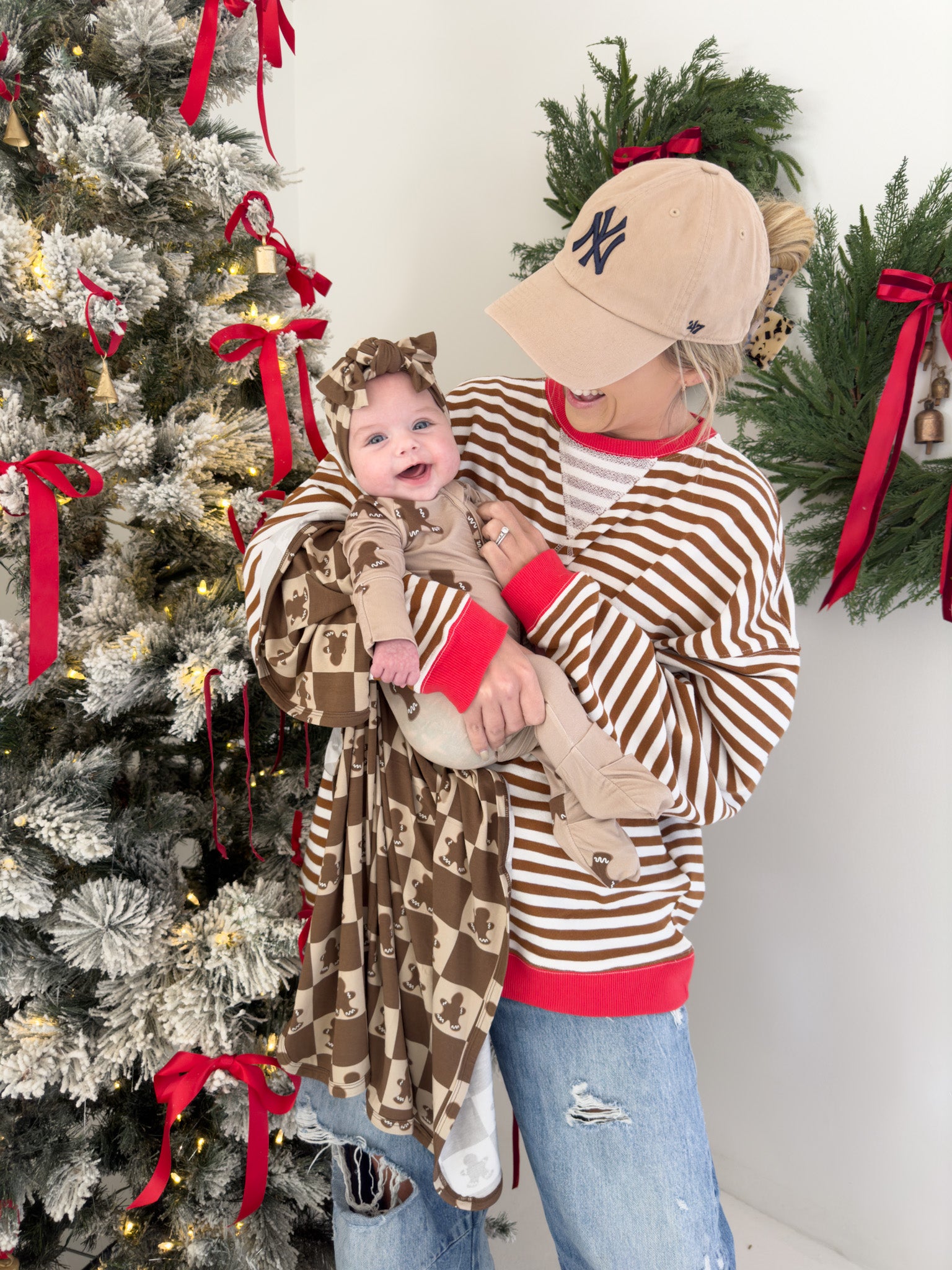 A woman in a beige cap and a brown and white striped sweater holds a smiling baby wrapped in the "Bamboo Swaddle | Gingerbread Checkerboard" by forever french baby. Dressed in matching outfits, they stand beside a Christmas tree adorned with red ribbons, surrounded by festive wreaths, enjoying the comfort of hypo-allergenic, breathable fabric.