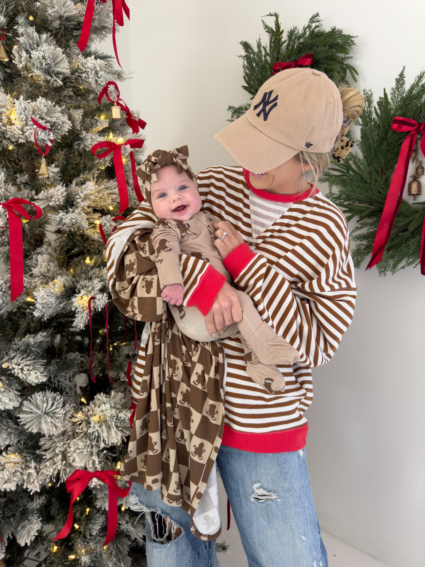 A person wearing a beige cap and striped sweater holds a smiling baby dressed in the Forever French Baby Bamboo Swaddle in the Gingerbread Checkerboard design, complete with a matching headband. They are standing by a decorated Christmas tree adorned with red ribbons, and there's a green wreath hanging on the wall.