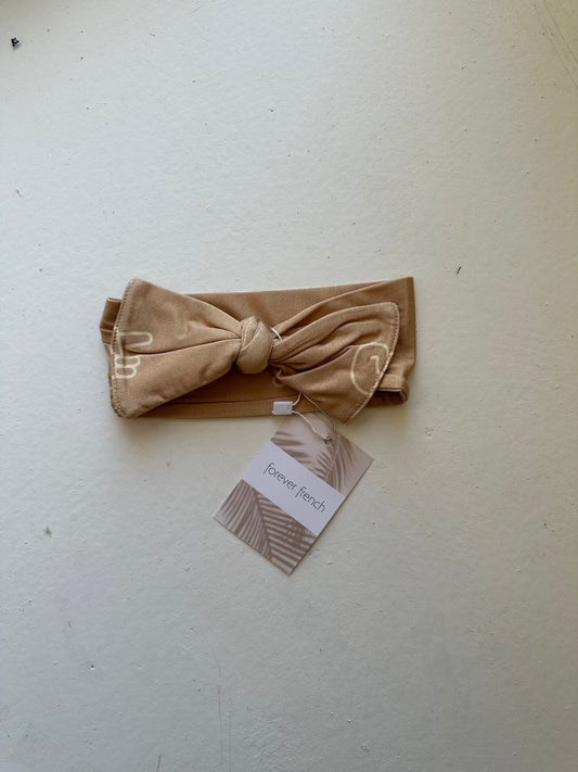 A beige Bamboo Head Wrap with a bow design sits on a light surface, featuring an abstract striped "forever french baby" tag.