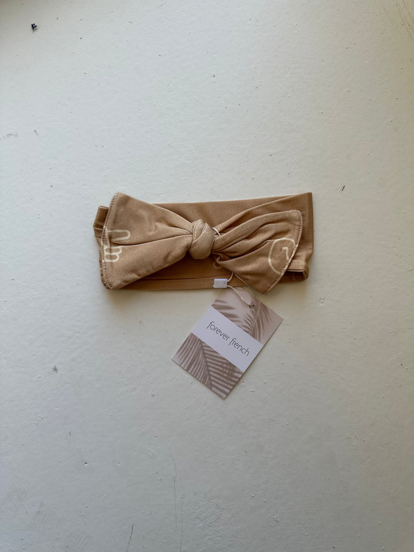 A beige headband with a large bow and knot detail rests on a light surface, featuring a small tag indicating the brand "forever french baby." The product name is Bamboo Head Wrap | Peace Sign.