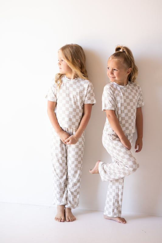 Two young girls wear the forever french baby Child Boxy Tee Lounge Set | Checkerboard, standing against a white wall—one barefoot with folded hands looking left, the other balancing on one foot, smiling to the right.