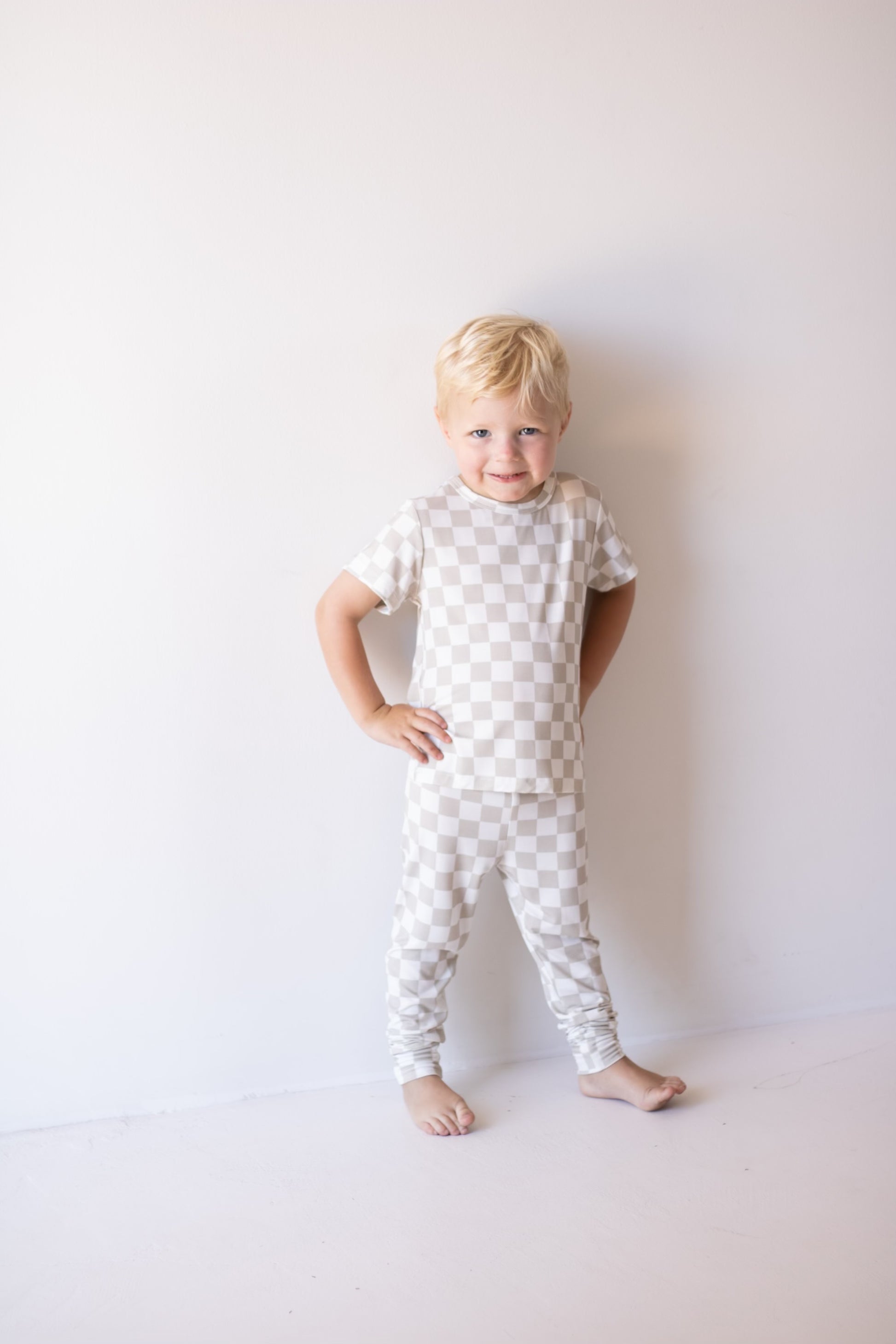 A young blond child stands barefoot against a white wall, smiling with one hand on their hip, wearing the forever french baby Bamboo Two Piece Lounge Set | Checkerboard.