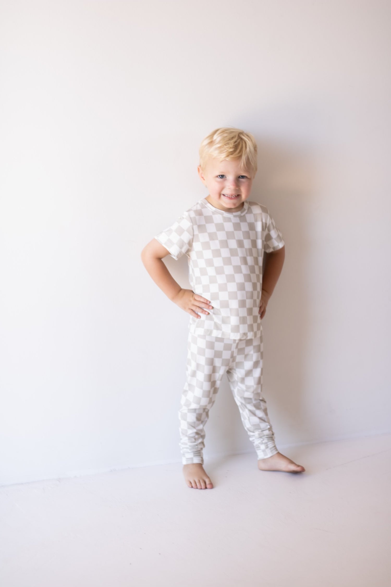 A young blonde child stands barefoot against a white wall, smiling with hands on hips, wearing the forever french baby Bamboo Two Piece Lounge Set | Checkerboard—hypoallergenic kids pajamas in neutral tones.