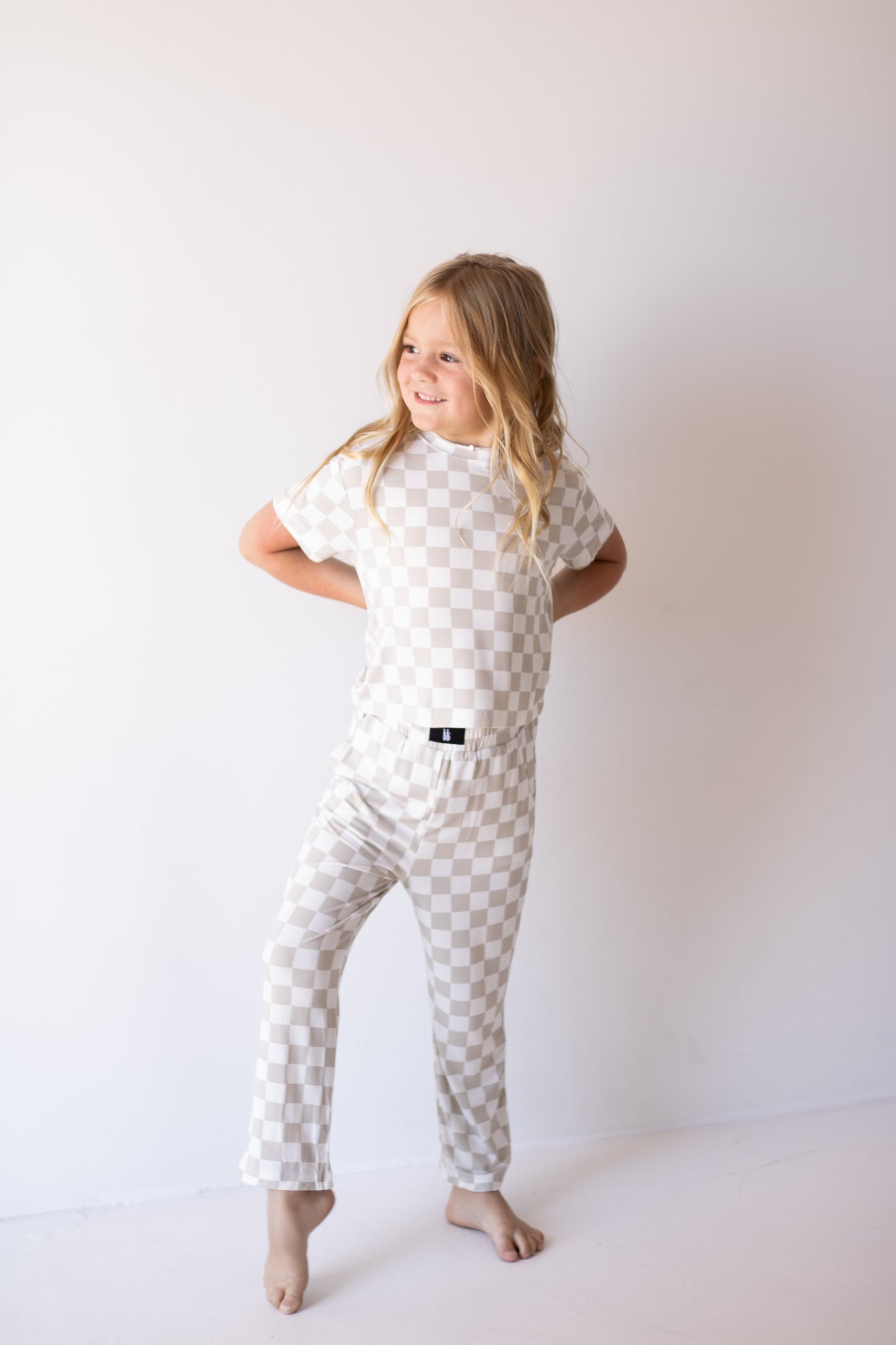 A young child with long blonde hair stands barefoot against a plain white background, smiling in breathable comfort while wearing the forever french baby Child Boxy Tee Lounge Set | Checkerboard.