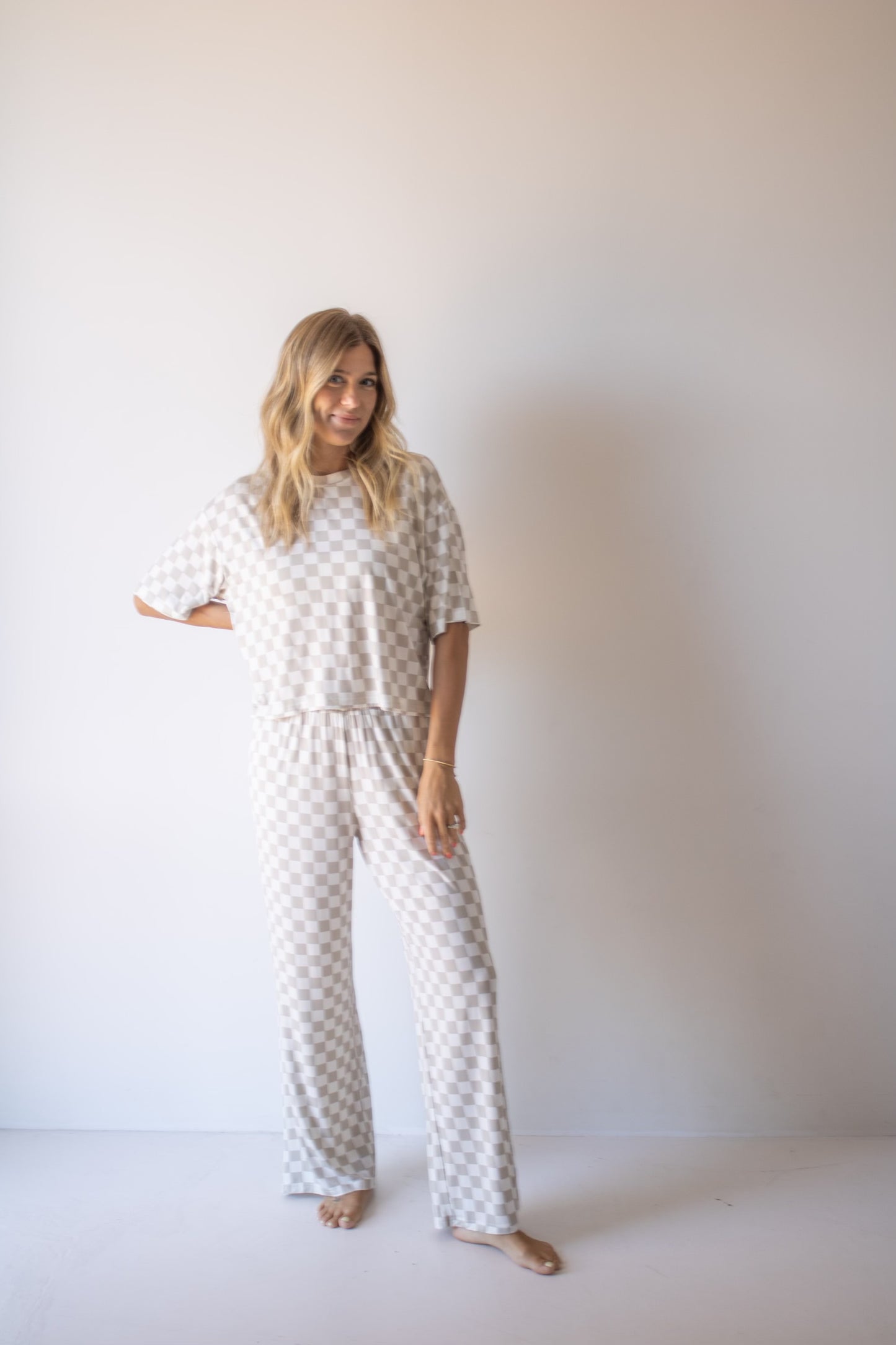 A woman with long blonde hair stands barefoot against a plain wall, smiling and posing in the forever french baby Women's Boxy Tee Lounge Set | Checkerboard—an effortlessly cozy loungewear look with wide pants and short sleeves.