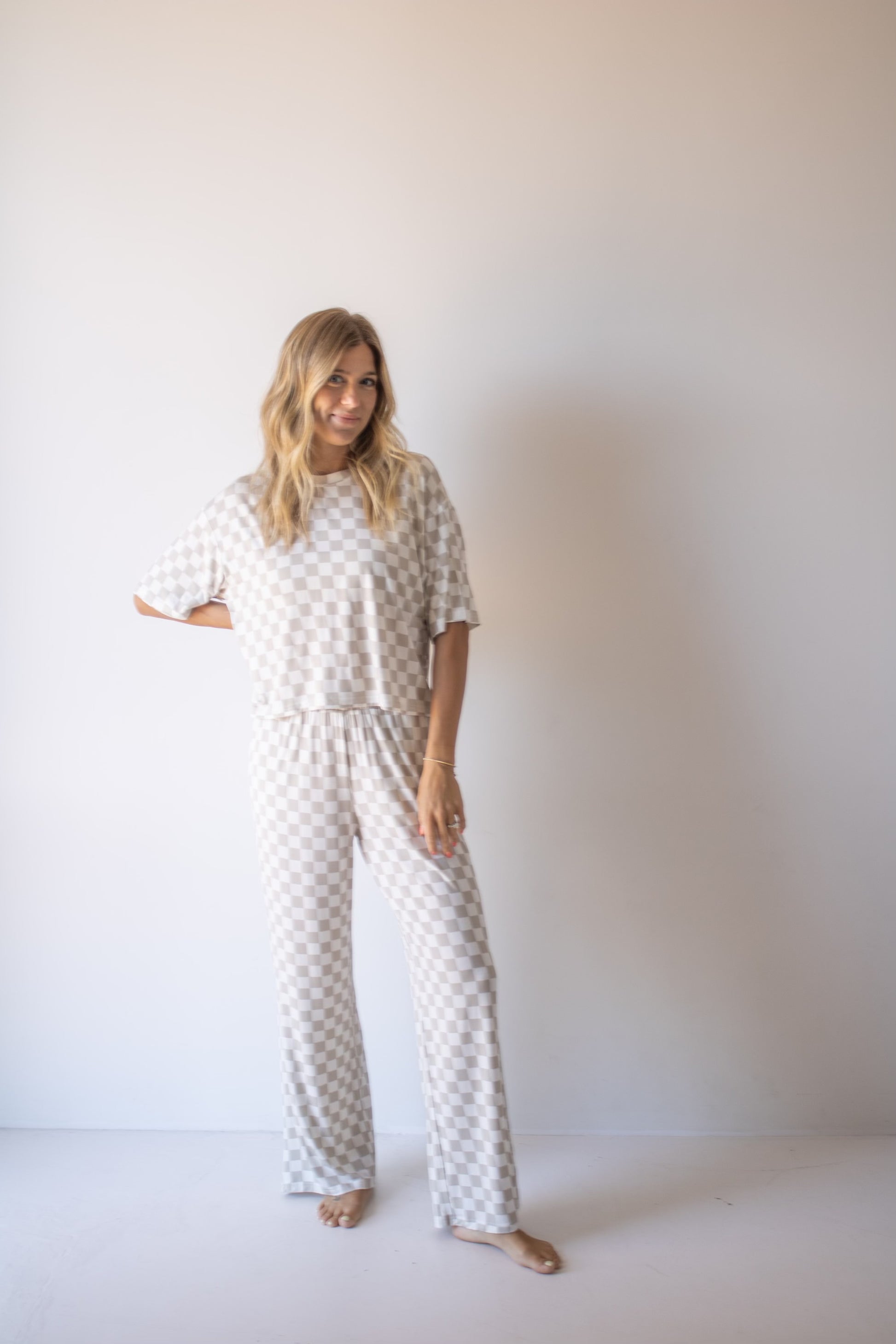 A woman with long blonde hair stands barefoot against a plain wall, smiling and posing in the forever french baby Women's Boxy Tee Lounge Set | Checkerboard—an effortlessly cozy loungewear look with wide pants and short sleeves.