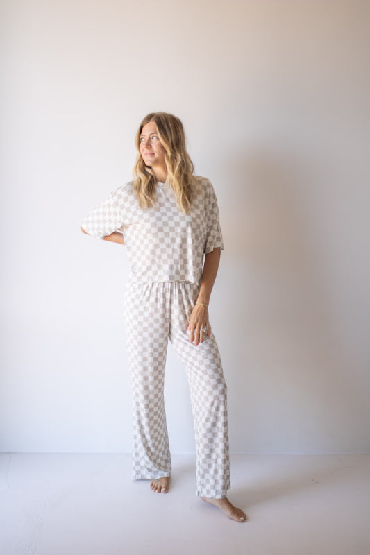 A woman stands barefoot against a plain white wall, smiling to her left with one hand on her hip. She wears the forever french baby Women's Boxy Tee Lounge Set in Checkerboard—ideal for stylish loungewear at home.