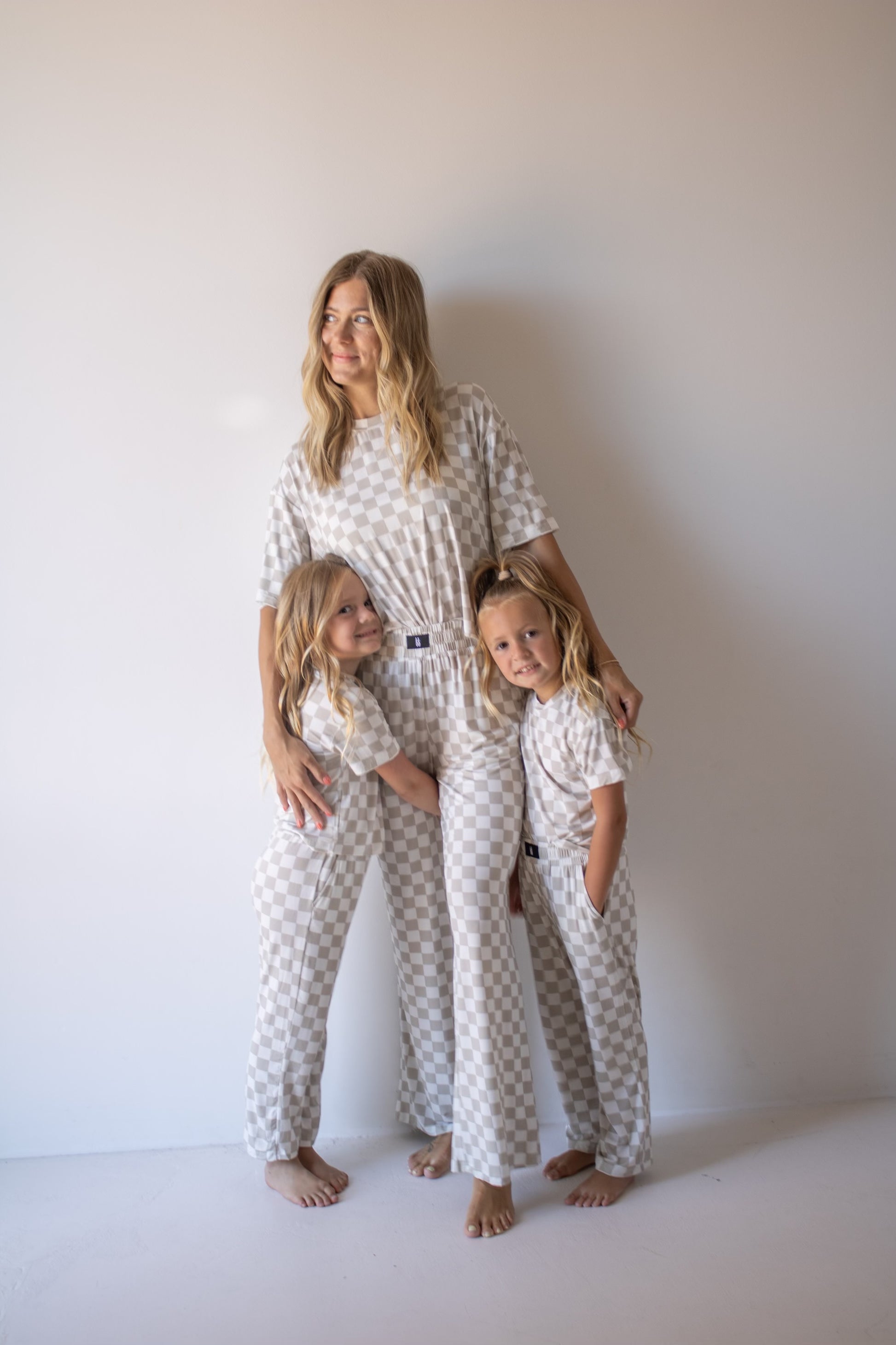 A woman and two young girls stand together against a plain wall, all looking content and relaxed in matching forever french baby Women's Boxy Tee Lounge Set | Checkerboard pajamas. The girls hug the woman, showing their close bond.
