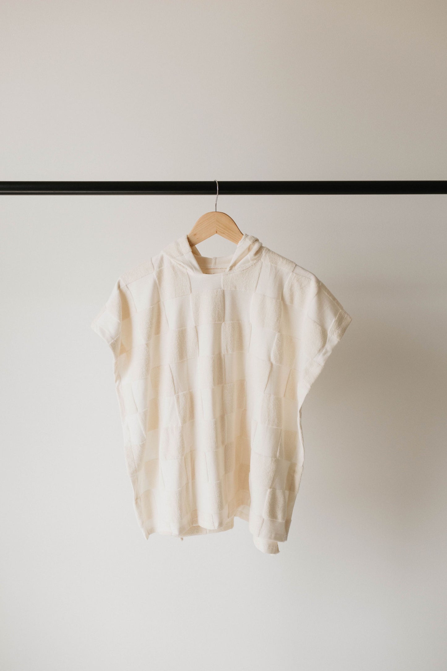 A cream-colored knit top with a textured checkerboard pattern is hanging on a wooden hanger against a plain white background. The top features short sleeves, a relaxed fit, and a round neckline, reminiscent of the effortless style from Forever French Baby.