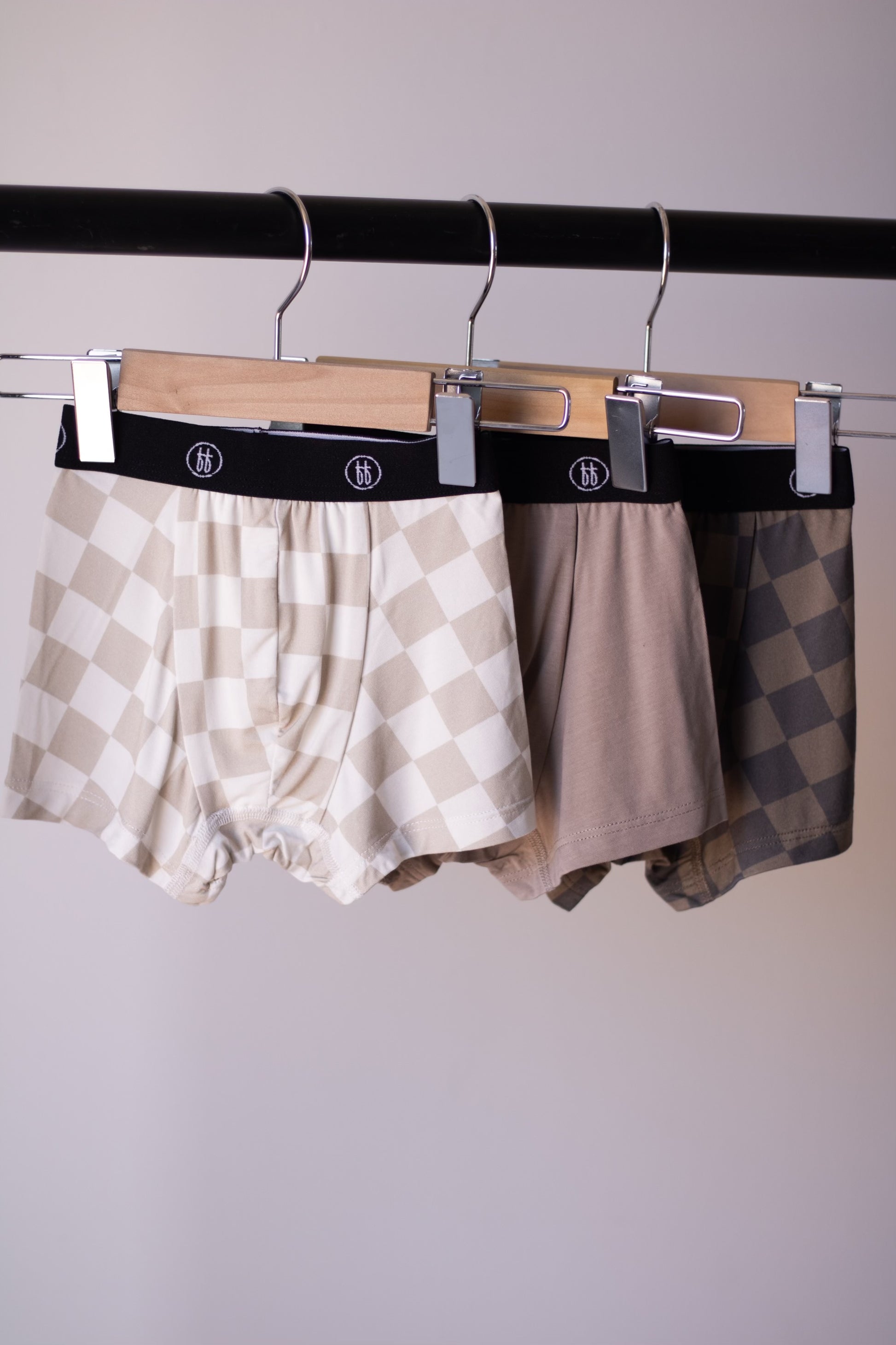 Three pairs of Bamboo Boxers (3 Pack) by forever french baby—Checkerboard and Faded Brown Checkerboard—hang from wooden clip hangers on a black rack. Enjoy hypo-allergenic comfort in stylish, neutral tones.