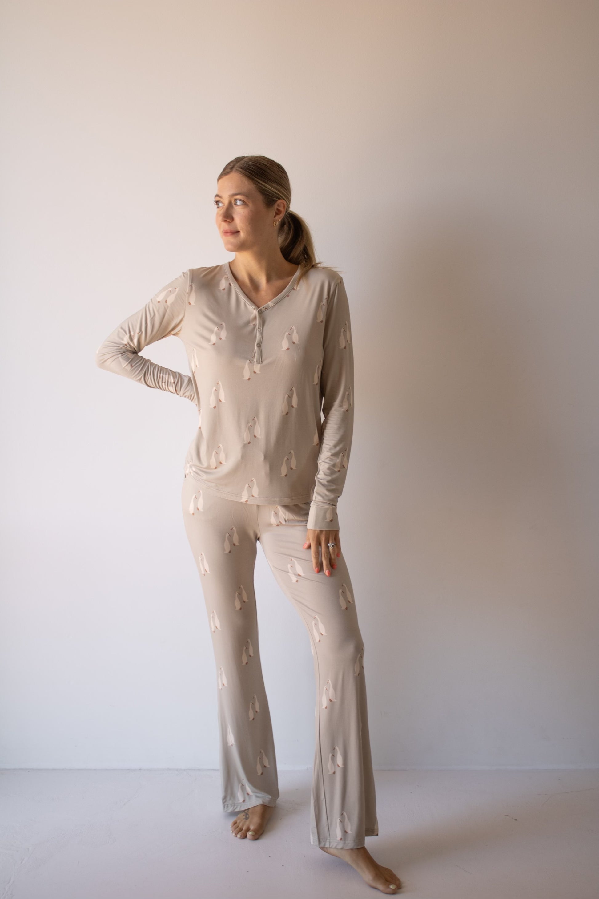 A woman stands barefoot against a plain wall, wearing the forever french baby Women's Bamboo Pajama in Silly Goose—a light beige set with a subtle print. She looks to the side, one hand on her hip and the other relaxed by her side.