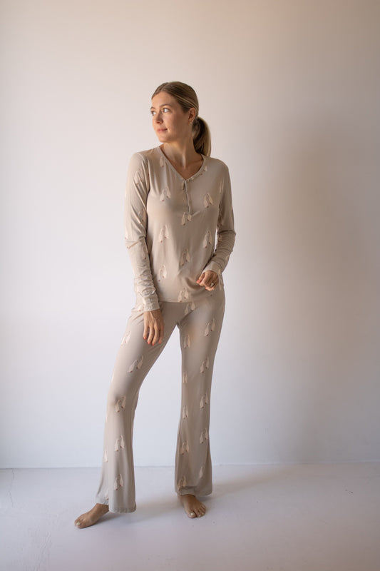 A woman stands barefoot against a plain wall, wearing forever french baby Women's Bamboo Pajama | Silly Goose with a subtle abstract pattern. She looks relaxed, gazing to the side with one hand crossed over the other.