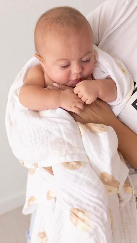 A baby wrapped in the forever french baby Muslin Swaddle in Tan & White Yin Yang patterns is being cradled by an adult. The baby looks down while holding part of the blanket close. The adult, dressed in a white shirt, gently holds the baby against their chest, creating timeless forever french baby moments.