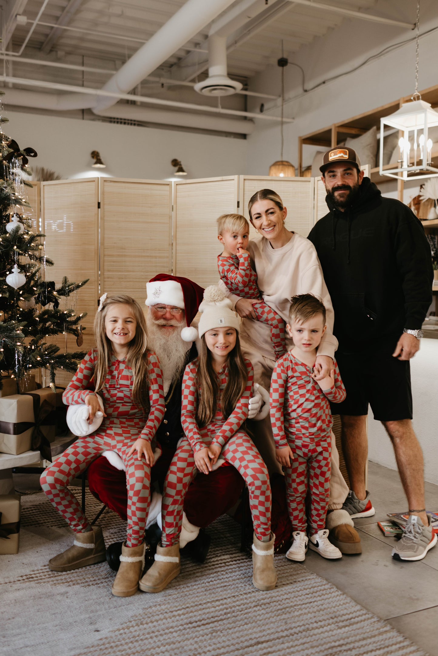 A family of six poses for a holiday photo with Santa Claus, beautifully captured by Amanda Riley Photos. Three children—two girls and one boy—wear matching red and gray pajamas as they stand in front of Santa. The mother holds a toddler dressed in Forever French Baby attire, while the father stands beside them. A decorated Christmas tree is on the left, completing this charming scene from the Santa Photos | Forever French Baby x Amanda Riley Photos collaboration.