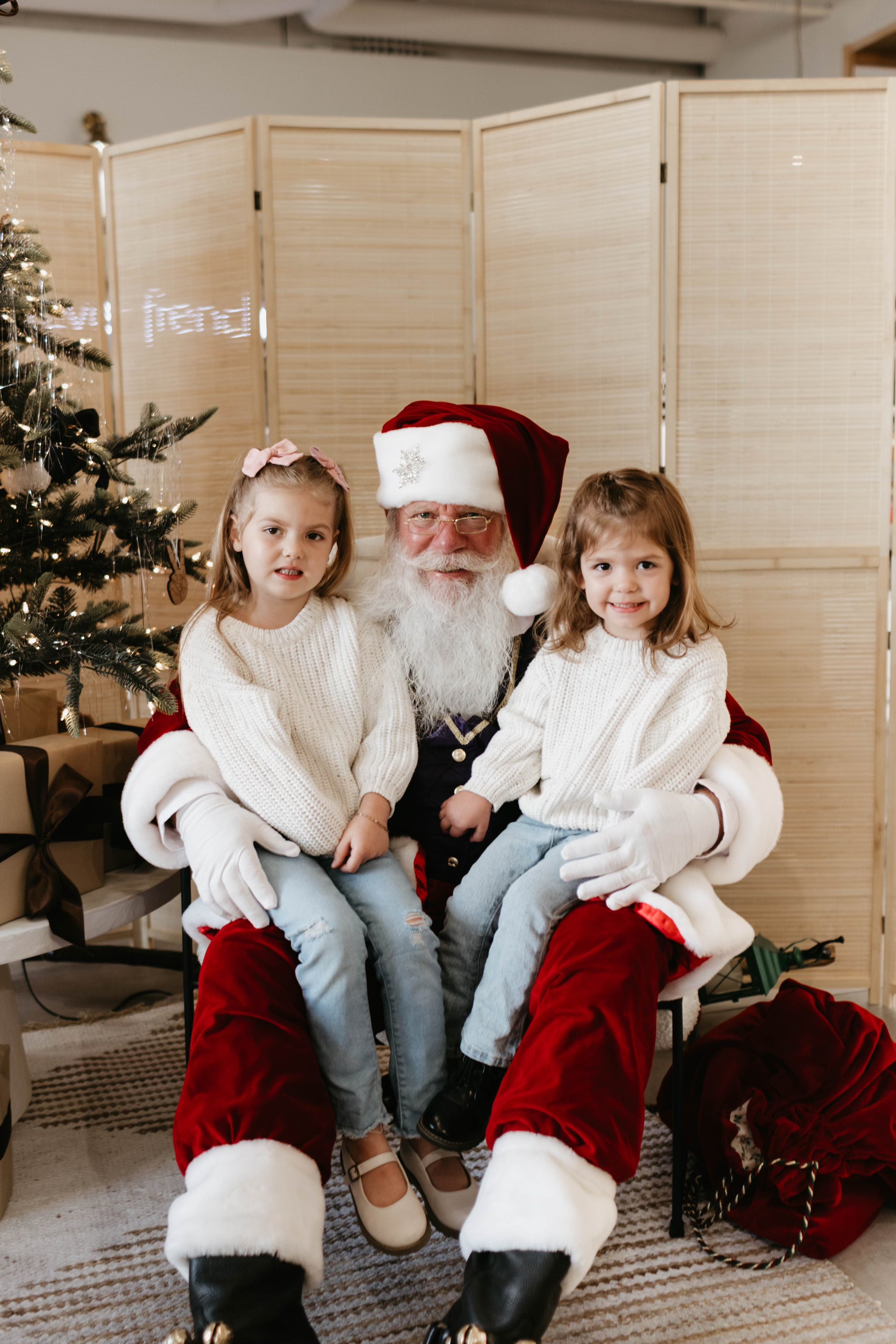 Two young children sit on Santa Claus's lap, both wearing white sweaters and light blue jeans. Santa is dressed in his traditional red and white suit. They are indoors, beside a decorated Christmas tree with presents underneath, captured perfectly by Amanda Riley Photos' "Santa Photos | Forever French Baby x Amanda Riley Photos," with a folding screen in the background.