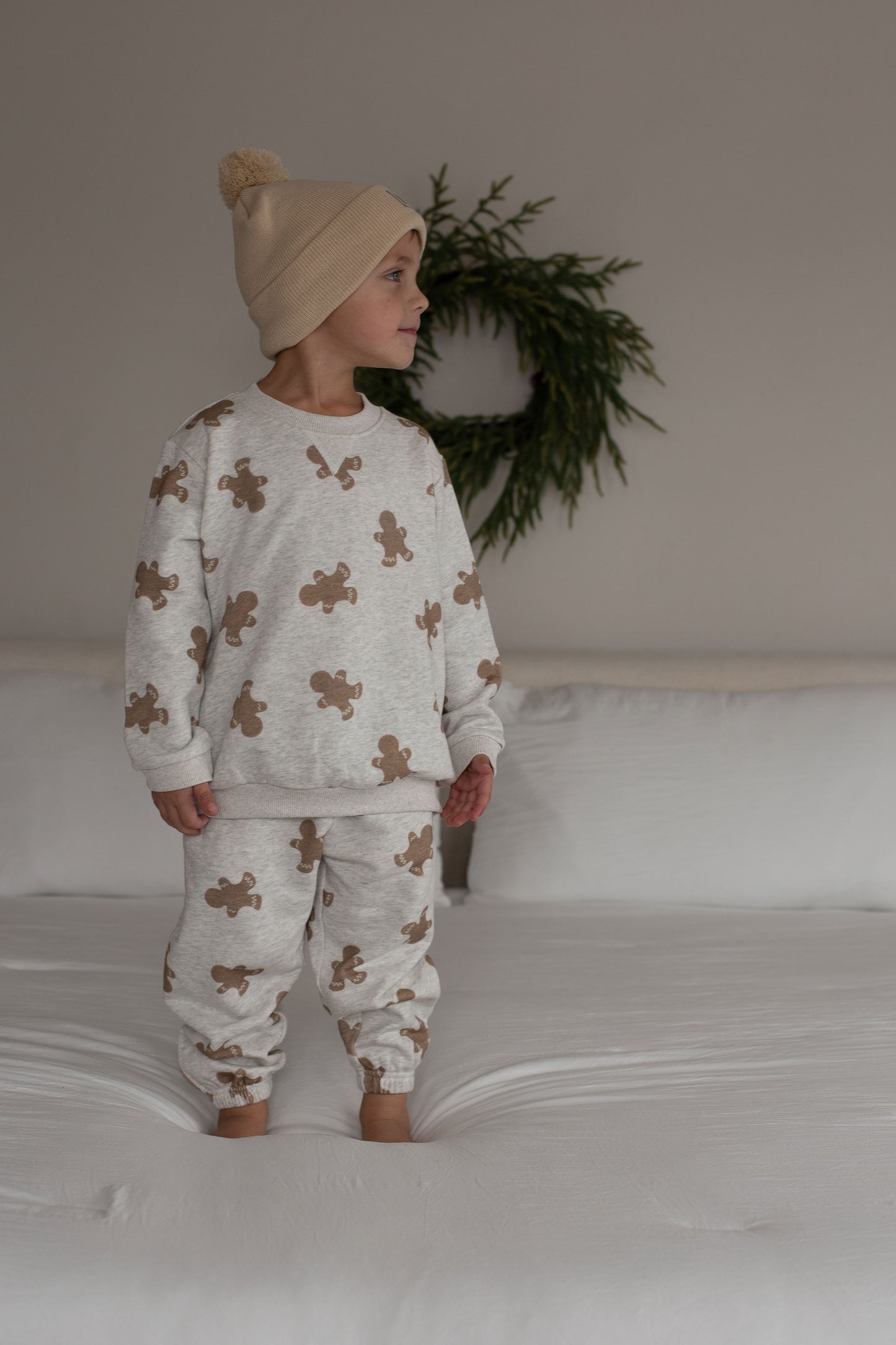 A young child in the forever french baby Sweat Set | Gingerbread 2.0 stands barefoot on a white bed, ready for everyday adventures. A green wreath decorates the neutral wall behind them.