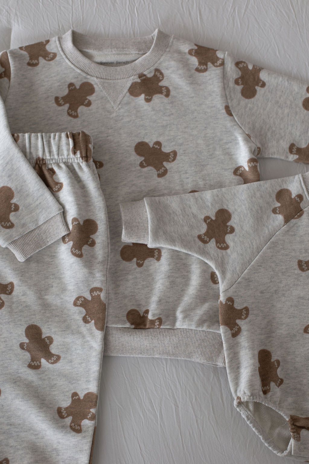 The forever french baby Sweat Set | Gingerbread 2.0 includes a light gray sweatshirt, pants, and onesie with a gingerbread print, perfect for everyday adventures and shown on a white textured background.