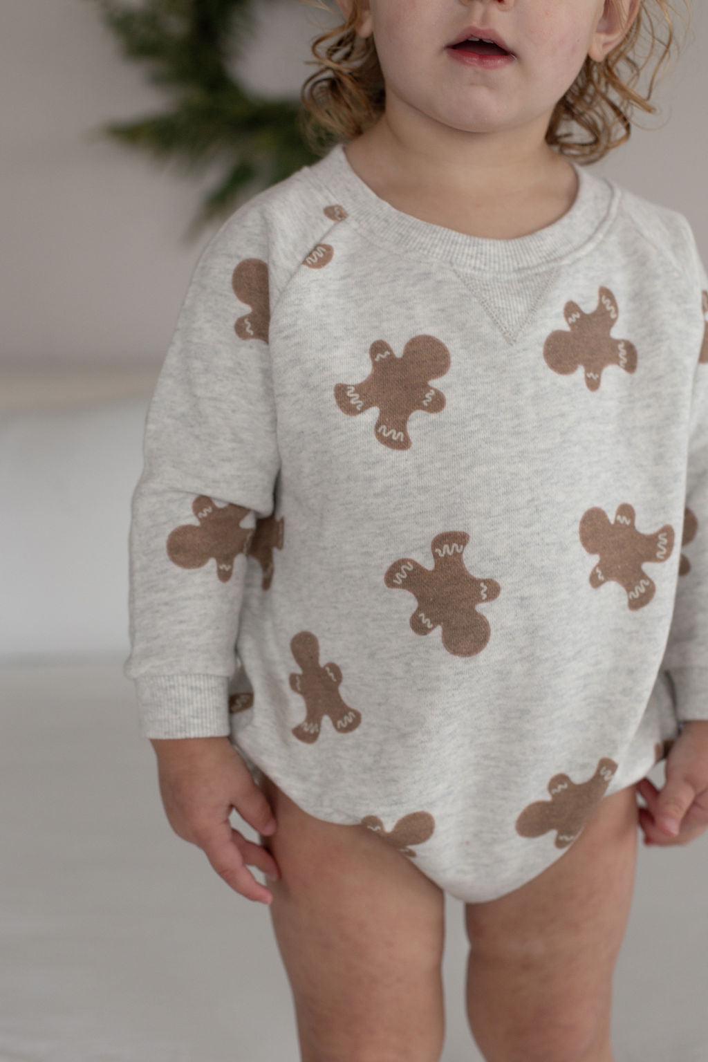 A young child stands indoors wearing the forever french baby Sweatshirt Romper | Gingerbread 2.0, a light gray romper patterned with brown gingerbread men. The child's face is partially out of frame and their mouth is slightly open.