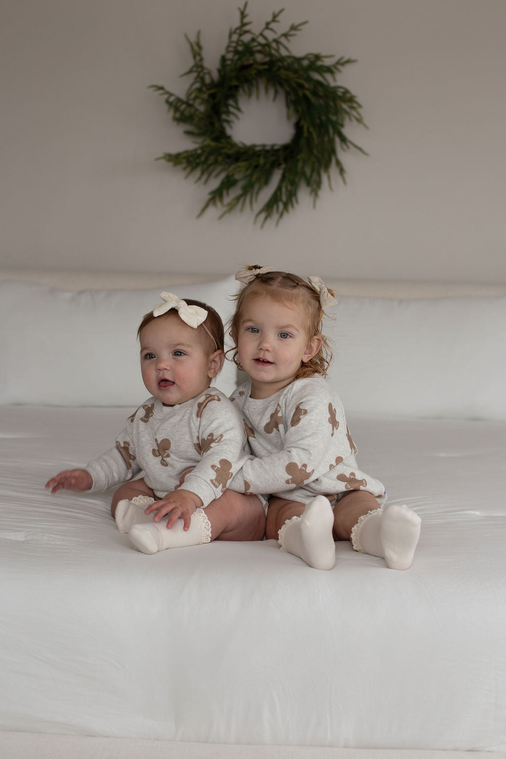 Two young children sit on a white bed, wearing matching forever french baby Sweatshirt Romper | Gingerbread 2.0 sets with brown floral patterns and white bows in their hair. A green wreath hangs on the wall behind them.