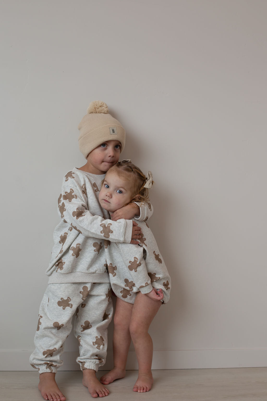 Two young children stand against a plain wall. The older one wears the forever french baby Sweat Set | Gingerbread 2.0 and hugs the younger, dressed in a matching outfit. Both barefoot, they're ready for everyday adventures together.