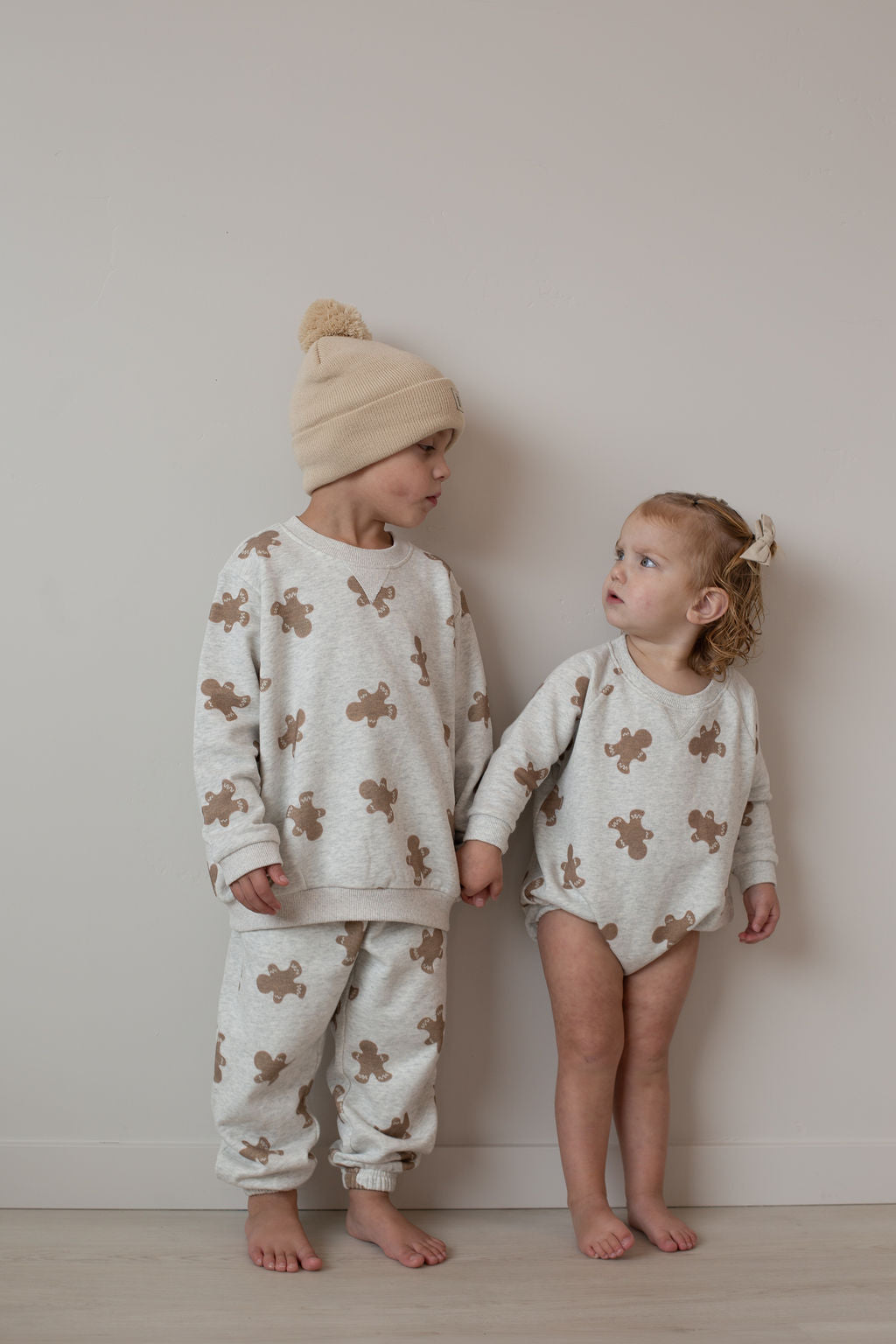 Two children in forever french baby’s Sweat Set | Gingerbread 2.0 hold hands by a plain wall, dressed in coordinating cream outfits with a sweet gingerbread print—the older in hat and pants, the younger in a bow and long-sleeve romper.