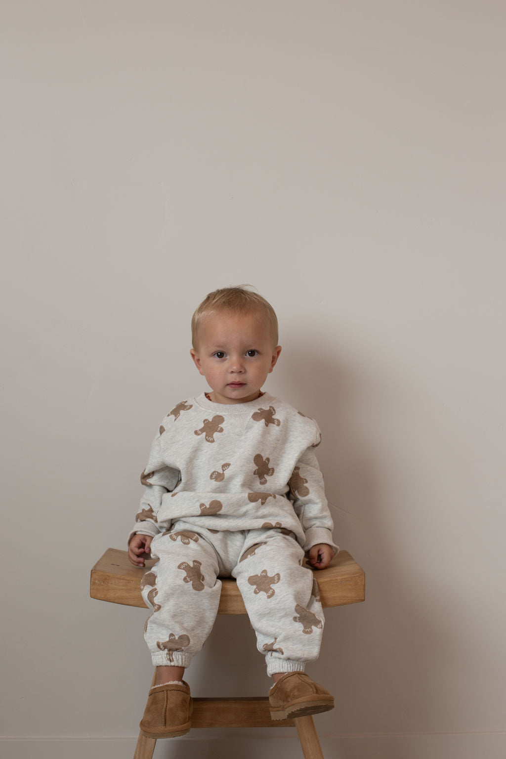 A young child with short blonde hair sits on a wooden stool against a beige background, wearing the forever french baby Sweat Set | Gingerbread 2.0 with playful brown teddy bear prints—perfect for everyday adventures.