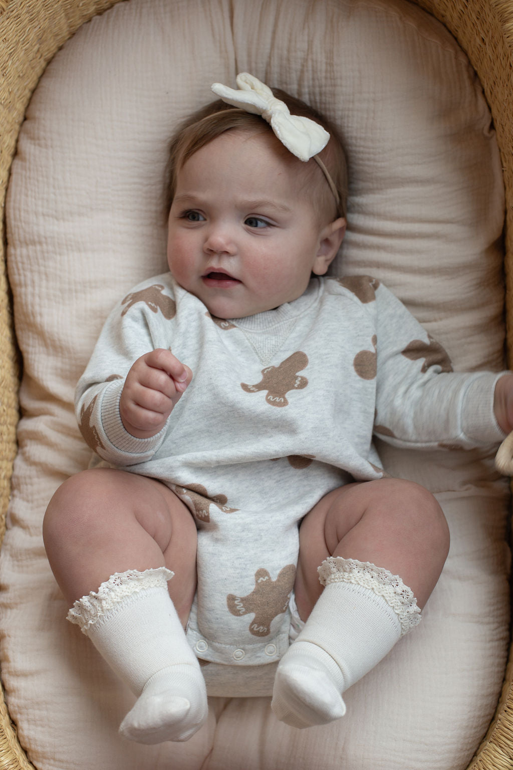 A baby wearing the forever french baby Sweatshirt Romper | Gingerbread 2.0—featuring a cream headband and white socks—lies comfortably on a cushioned surface in a woven basket, gazing softly to the side.