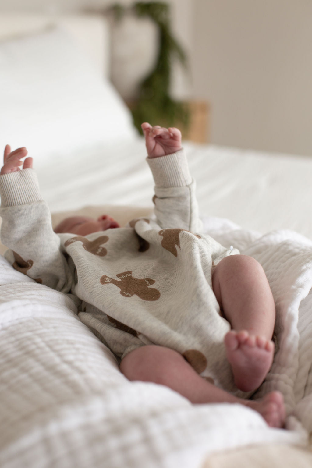 A baby lies on a white bed with arms raised, dressed in the forever french baby Sweatshirt Romper | Gingerbread 2.0—a cozy, light outfit with charming prints. The softly focused background adds an inviting touch.