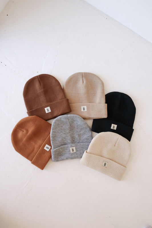 Six folded ff Knit Beanie | Adult hats from forever french baby are neatly laid out on a white surface. Made from breathable yarn, these hats come in a variety of colors: brown, light gray, black, tan, dark brown, and beige. Each beanie features a ribbed design and includes a small white tag with the brand's simple logo on the front.