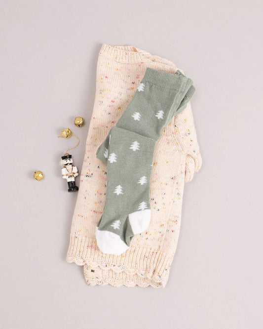 A folded cream knit sweater with colored specks is topped with Little Millers Pine Tights—sage green with white tree patterns and white toes. Nearby are three small gold bells and a white toy soldier figure.
