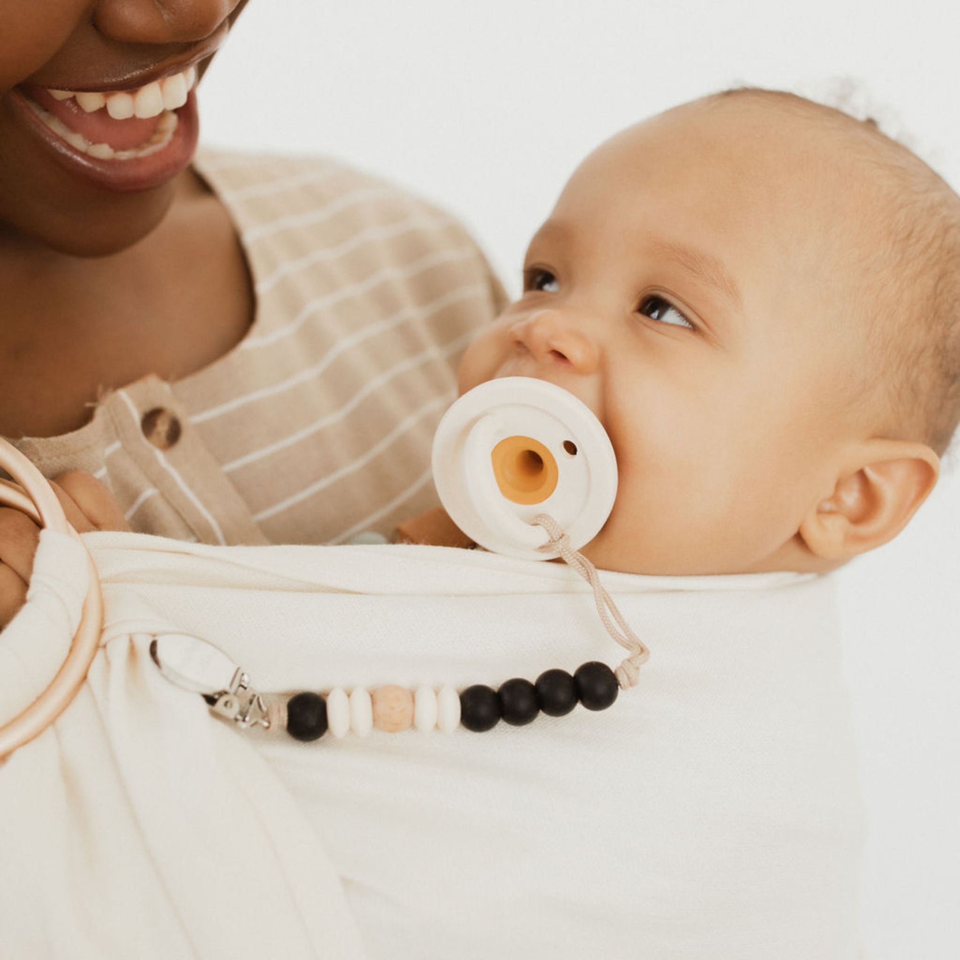 A close-up of a smiling woman holding a baby swaddled in a white blanket. The baby, using a Modern Pacifier in Caramel from forever french baby, is looking up at the woman. The woman is wearing a beige and white striped top, and the baby has a pacifier clip with black and beige beads.