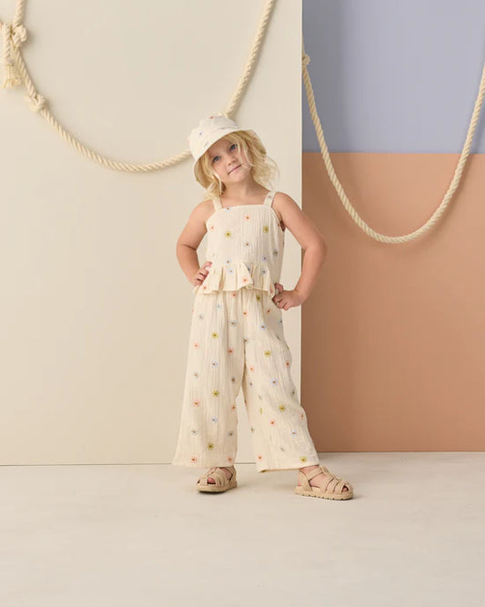 A young child models the Rylee + Cru Kayli Set | Embroidered Daisies—a cream-colored, cropped tank and wide leg pants adorned with embroidered daisies—paired with matching sandals and a bucket hat, posing confidently in front of a pastel geometric backdrop with thick ropes.