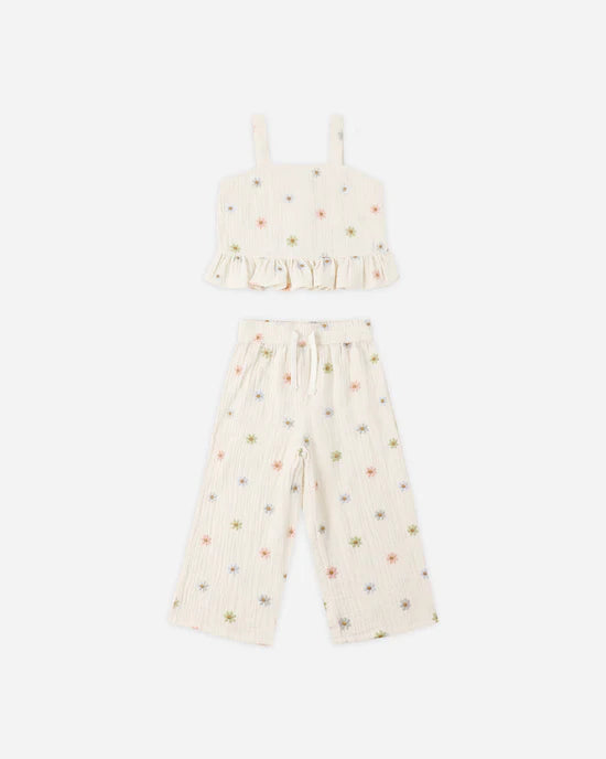 The Kayli Set by Rylee + Cru features a two-piece cream-colored outfit, including a sleeveless cropped tank with ruffled accents and wide-leg pants, both decorated with small, colorful embroidered daisies.