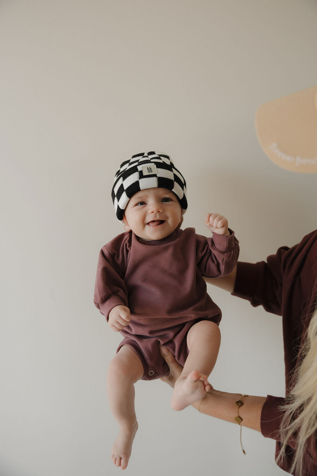 A smiling baby wears the forever french baby Sweatshirt Romper | Twilight in maroon and a black-and-white checkered beanie, held up by an adult with a plain light background.