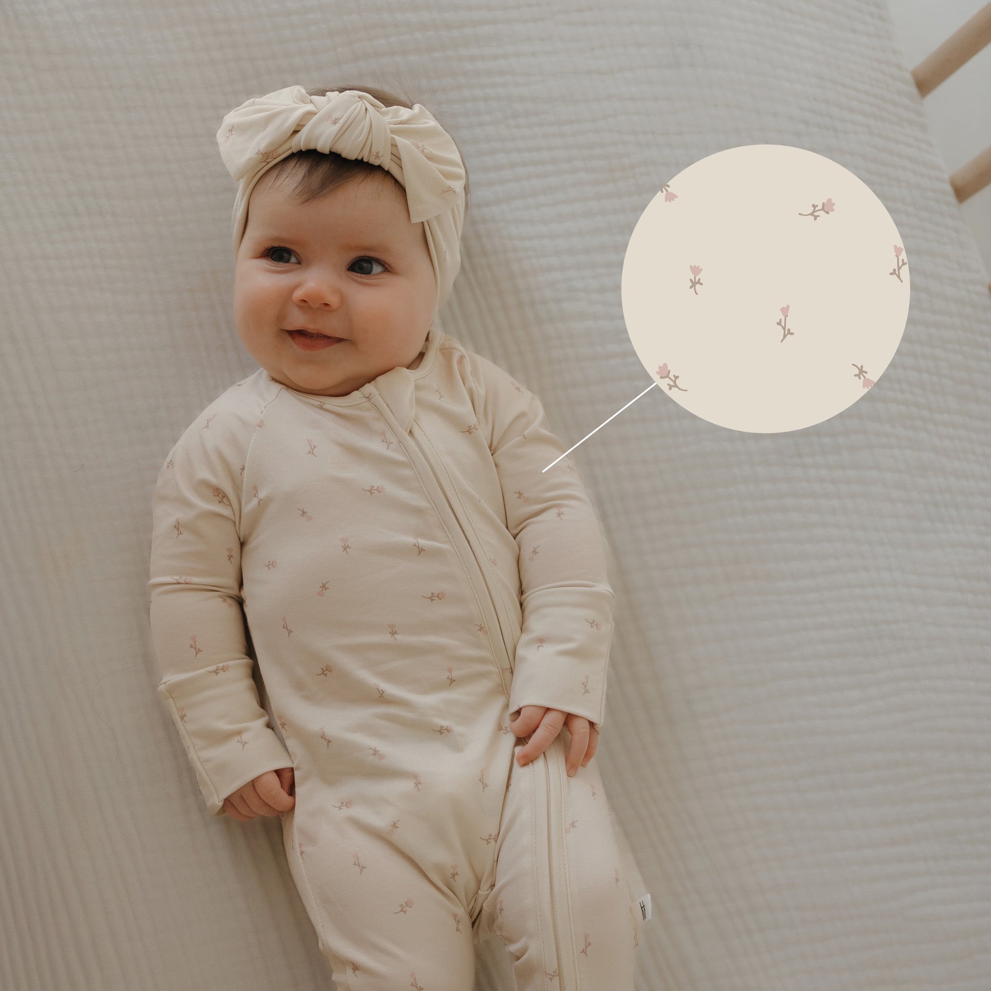 A baby in cream-colored, hypoallergenic Bamboo Zip Pajamas | Lucy from forever french baby, with a matching headband, lies on a textured white blanket. A zoomed-in circle shows the subtle pink floral pattern on the soft fabric.