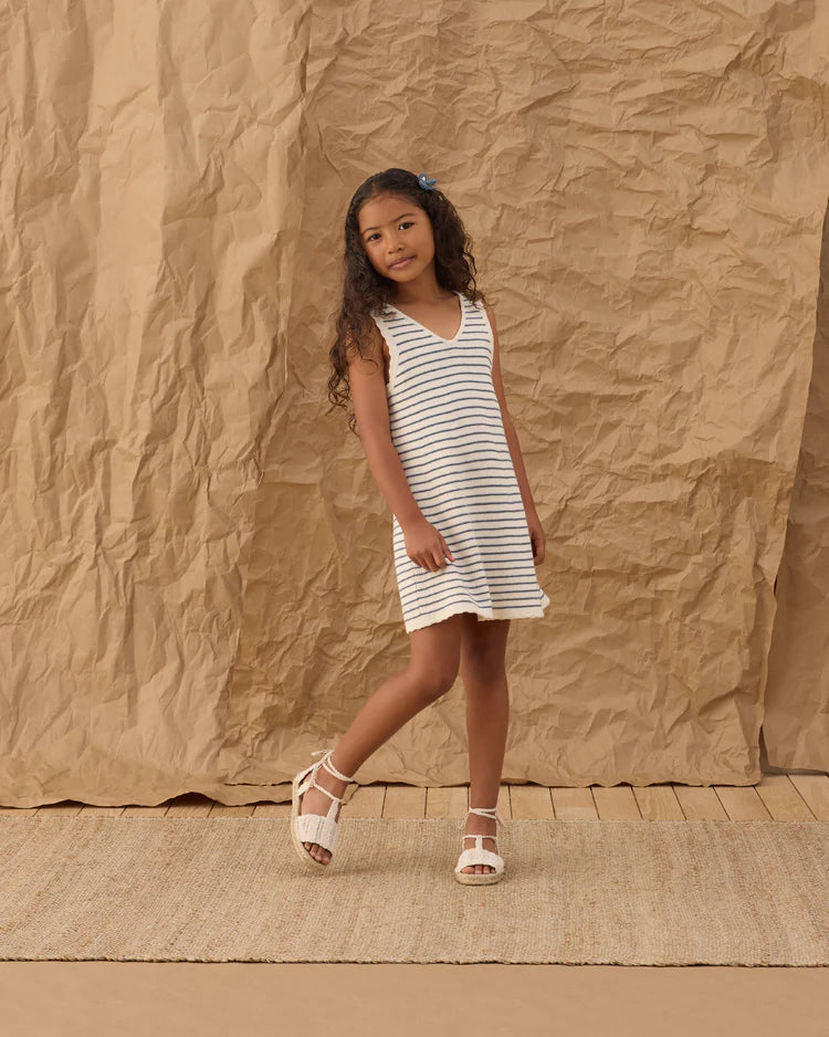 A young girl with long curly hair wears the Rylee + Cru Crochet Tank Mini Dress in Marine Stripe, paired with white sandals. She stands on a woven rug against a crumpled brown paper backdrop—a true summer staple look.