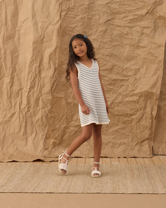A young girl with long curly hair wears the Rylee + Cru Crochet Tank Mini Dress in Marine Stripe, paired with white sandals. She stands on a woven rug against a crumpled brown paper backdrop—a true summer staple look.
