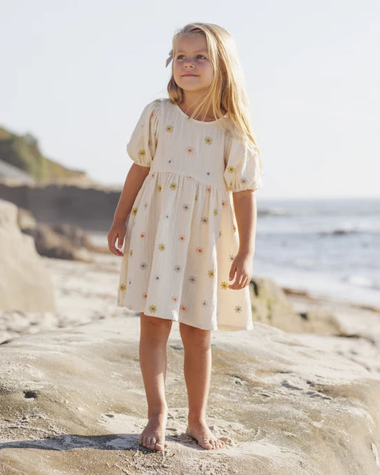 A young girl with long blonde hair stands barefoot on a rocky beach, wearing the Marley Dress | Embroidered Daisies by Rylee + Cru—a light-colored baby doll dress delicately decorated with small embroidered daisies. The ocean and shoreline are visible in the background on a sunny day.