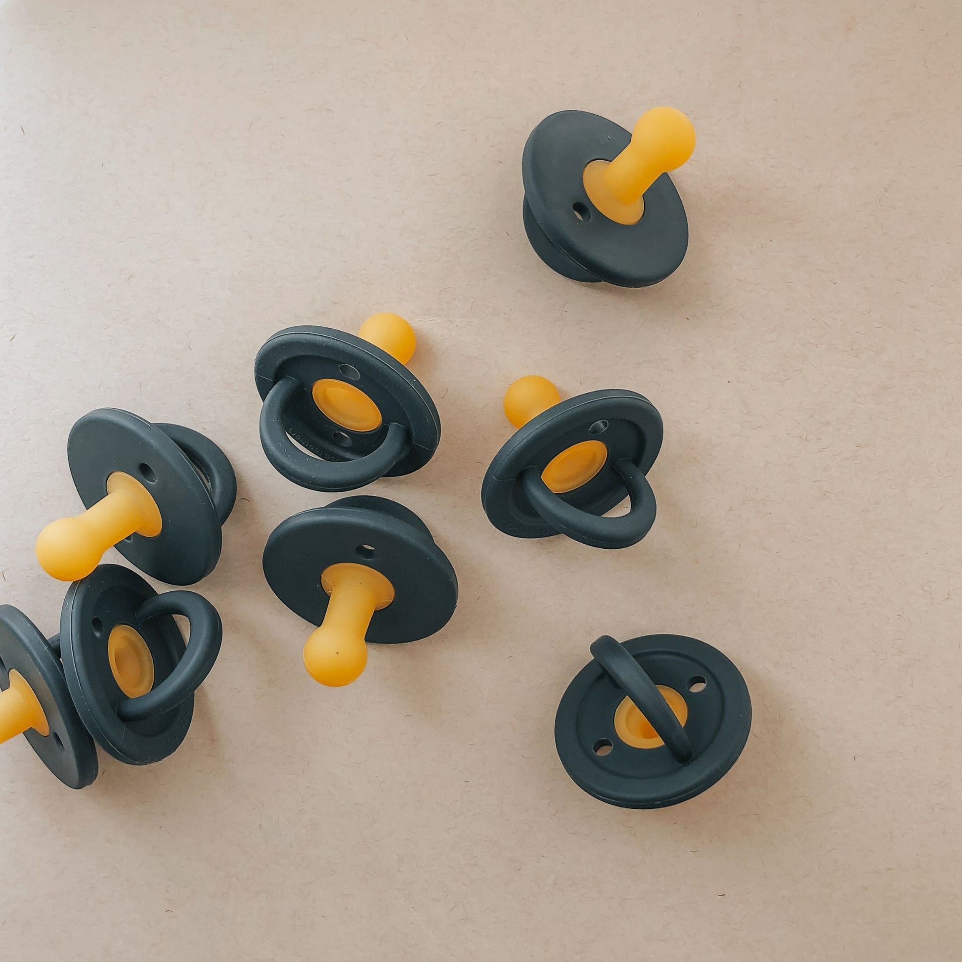 A group of Modern Pacifiers by forever french baby in navy are scattered on a light beige surface. These essential baby items feature round, navy shields with attached yellow nipples. They are arranged in a casual, unsystematic manner.