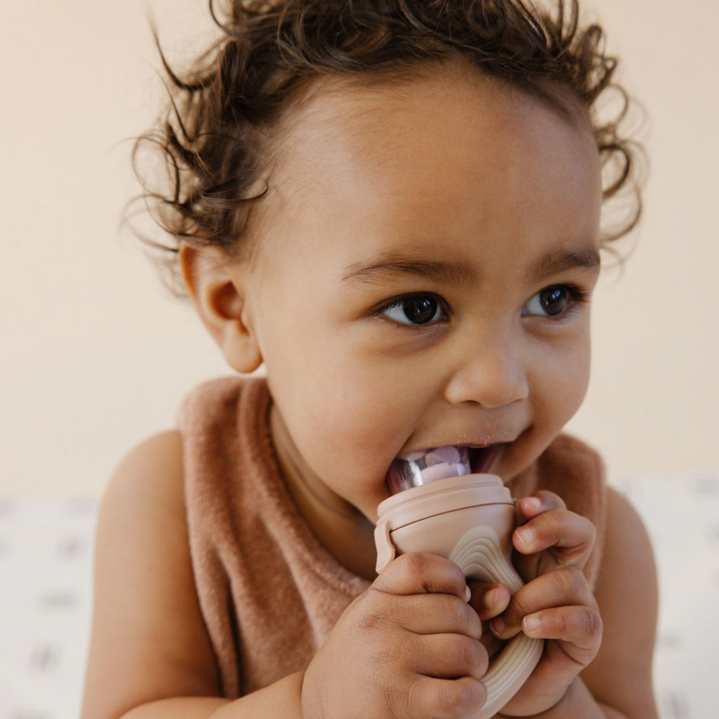 A young child with curly hair is holding a Modern Mod Feeder by forever french baby and chewing on it. The child is wearing a sleeveless, light brown shirt. The background is softly blurred, drawing focus to the child's expression and activity.