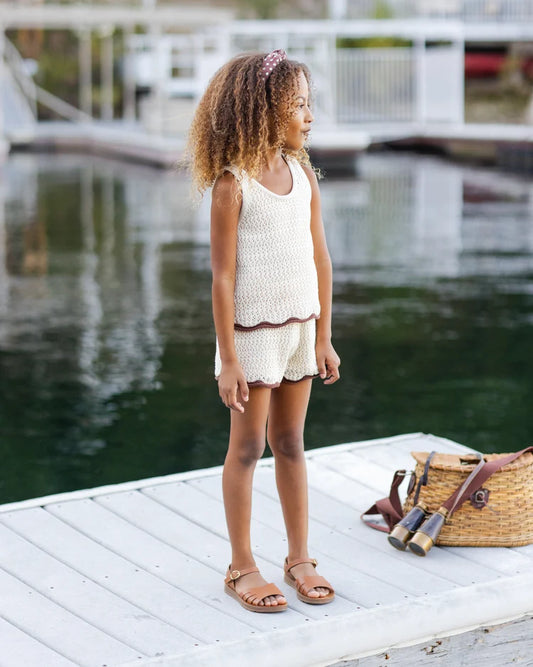 A young girl with curly hair stands on a dock by the water, wearing the Rylee + Cru Crochet Tank Set in Natural—her perfect summer staple—paired with brown sandals. A woven bag and binoculars rest on the dock beside her.