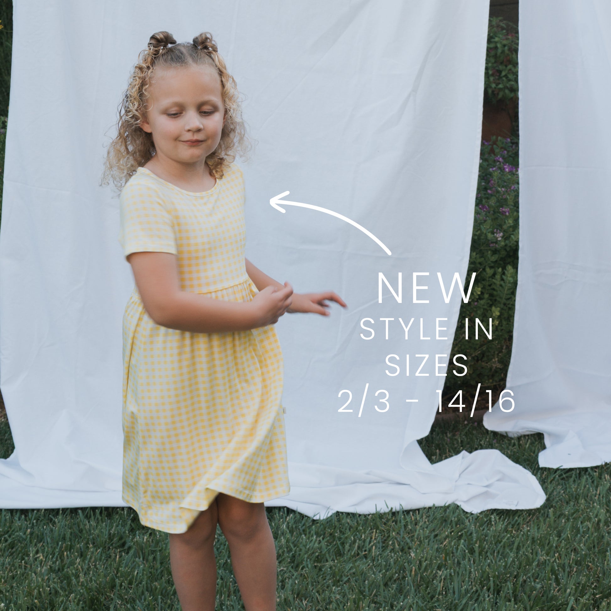 A young girl with curly blond hair wears the forever french baby Bamboo T-Shirt Dress in Citron and stands on grass before a white sheet. Text reads: "NEW style in sizes 2/3 – 14/16" with an arrow pointing to her dress.