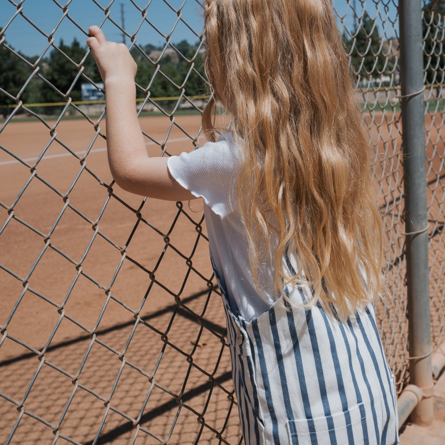 A young girl with long blonde hair, wearing a white shirt and forever french baby Overalls | Denim Stripe, stands facing a chain-link fence, gazing at an empty baseball field beneath a clear blue sky.
