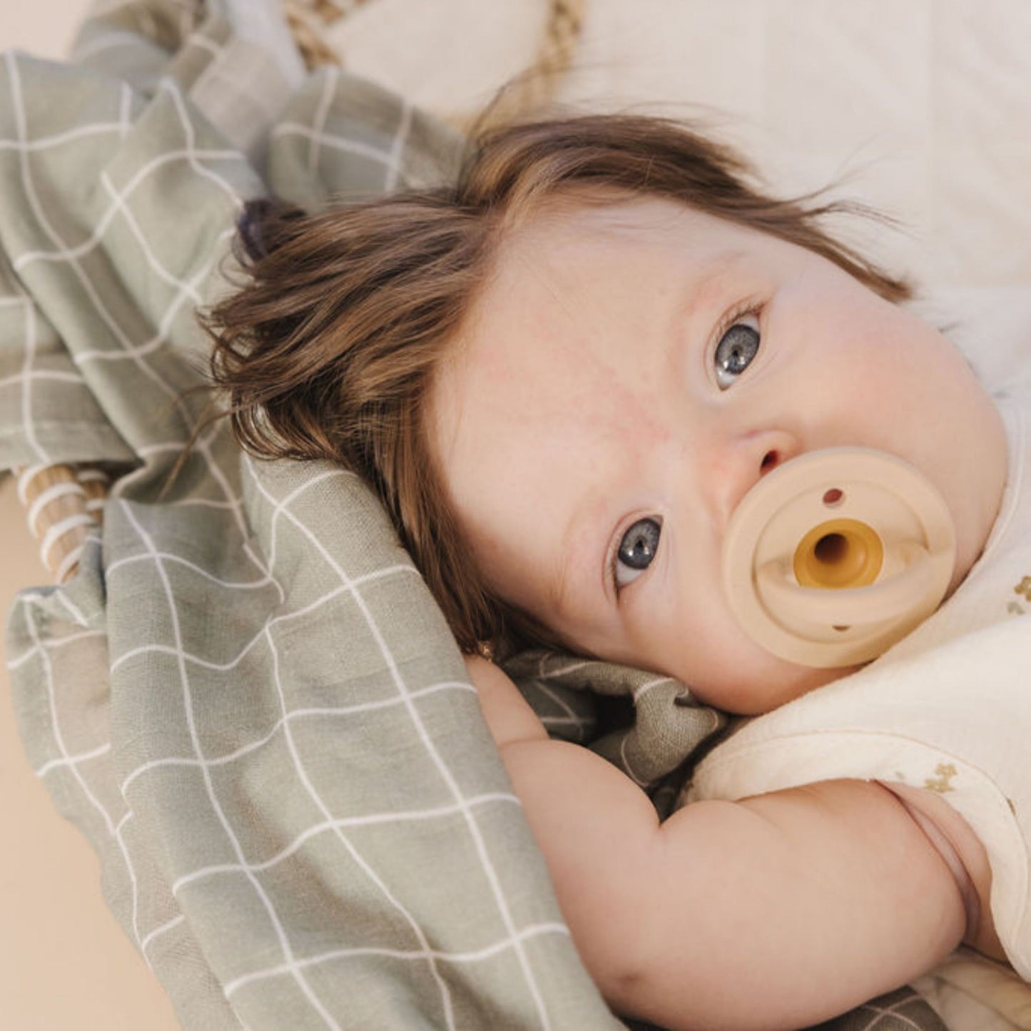 A baby with light brown hair lies on a checked blanket, looking up with blue-gray eyes. The baby is sucking on a Modern Pacifier | Fawn from forever french baby and wearing a white sleeveless outfit. The scene appears calm and serene, highlighting the importance of baby essentials in creating such peaceful moments.