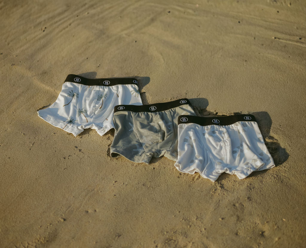 Three pairs of forever french baby Bamboo Boxers (3 Pack)—Lifesaver, Starfish & Palm Tree—featuring black waistbands are laid out on sand, offering ultra-soft bamboo comfort for kids with sensitive skin.