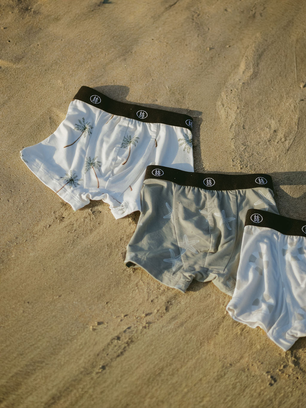 Three pairs of men's bamboo boxers by forever french baby lie on sand. Designs include Lifesaver, Starfish, and Palm Tree—each with a black waistband. These hypo-allergenic boxers offer ultimate comfort in a 3 pack set.