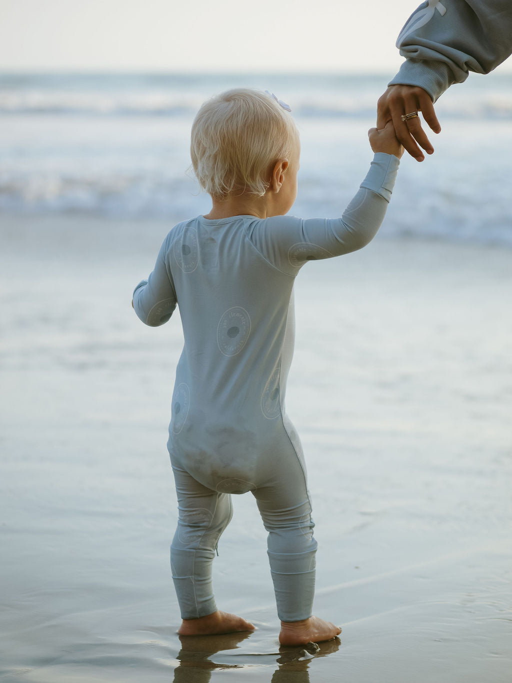 A small blond-haired child wearing forever french baby Bamboo Zip Pajamas | San Clemente stands on wet beach sand, holding an adult’s hand, with ocean waves in the background.